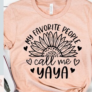 K&ouml;nnte beinhalten: Ein pfirsichfarbenes T-Shirt mit einer schwarzen Sonnenblumengrafik und dem Text "Meine Lieblingsmenschen nennen mich Yaya" mit Herzen.