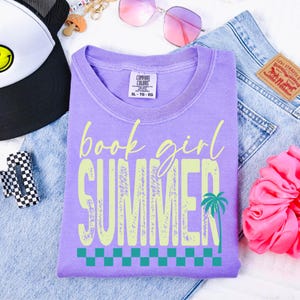 May include: A lilac Comfort Colors t-shirt with the words "book girl SUMMER" in a light green, distressed font. A palm tree graphic and a checkered pattern are below the text. The shirt is folded on top of denim jeans.