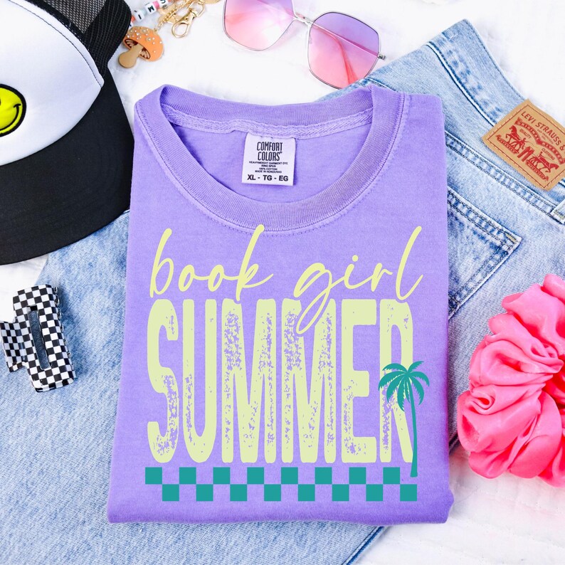 May include: A lilac Comfort Colors t-shirt with the words "book girl SUMMER" in a light green, distressed font. A palm tree graphic and a checkered pattern are below the text. The shirt is folded on top of denim jeans.