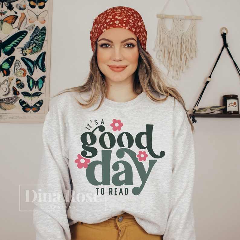 Op de afbeelding: Een wit sweatshirt met een groene en roze afbeelding waarop staat "It's a good day to read". De afbeelding is omringd door bloemen. Het model draagt een rood bloemenband.