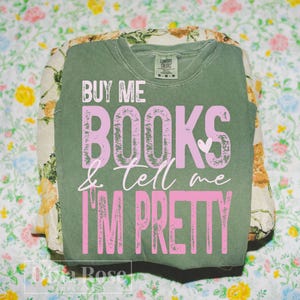 May include: An olive green t-shirt with the text "BUY ME BOOKS & TELL ME I'M PRETTY" in pink. The shirt is folded and rests on top of a floral patterned fabric. The shirt has a crew neck and short sleeves.