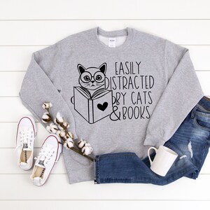 K&ouml;nnte beinhalten: Grauer Pullover mit einer schwarz-wei&szlig;en Illustration einer Katze, die ein Buch liest. Der Text auf dem Pullover lautet "Easily Distracted by Cats & Books".