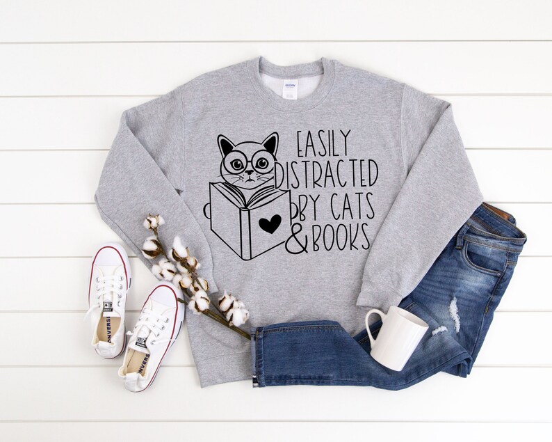 K&ouml;nnte beinhalten: Grauer Pullover mit einer schwarz-wei&szlig;en Illustration einer Katze, die ein Buch liest. Der Text auf dem Pullover lautet "Easily Distracted by Cats & Books".