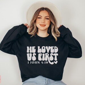 May include: Black sweatshirt with white text that reads "He Loved Us First 1 John 4:19" in a retro font. The sweatshirt is being worn by a person with long brown hair and a white hat.