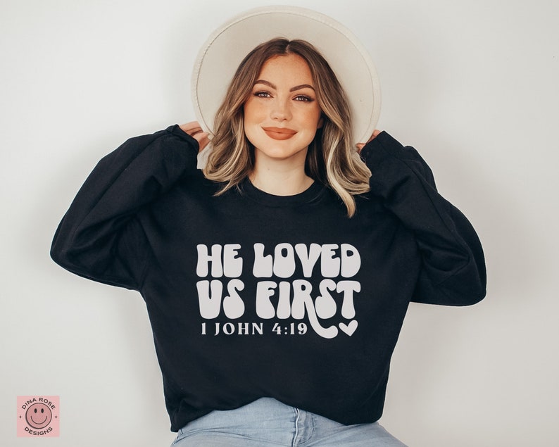 May include: Black sweatshirt with white text that reads "He Loved Us First 1 John 4:19" in a retro font. The sweatshirt is being worn by a person with long brown hair and a white hat.