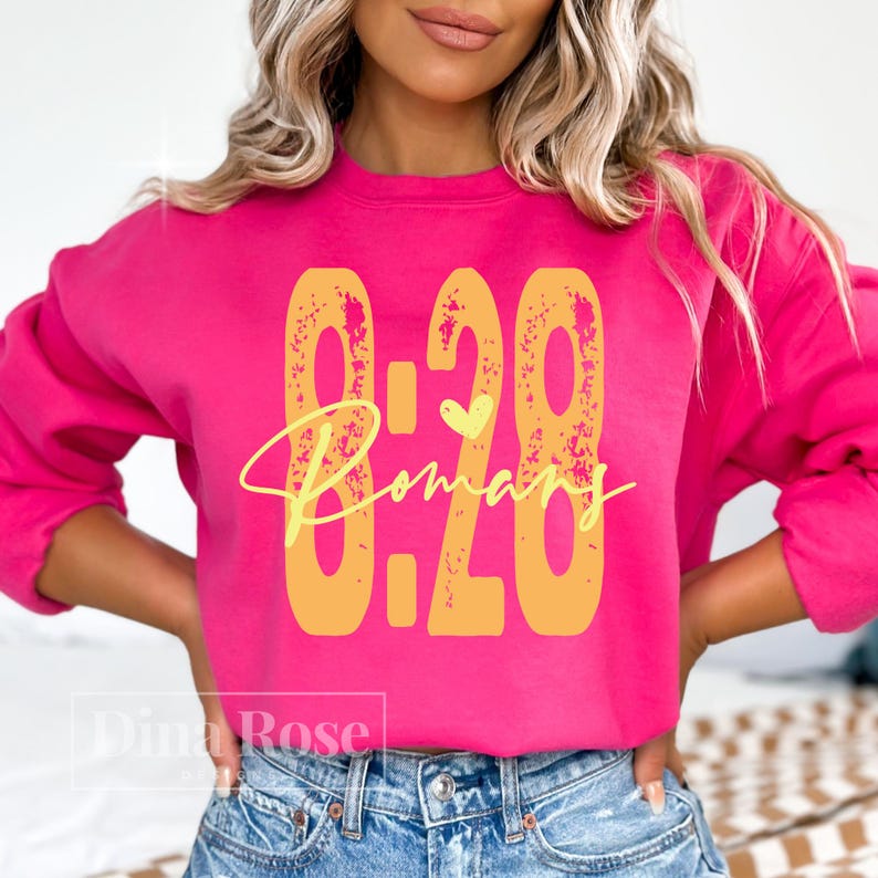 May include: A bright pink sweatshirt with the text "8:28 Romans" in yellow. The numbers and text have a distressed, textured appearance. A small yellow heart is above the word "Romans".