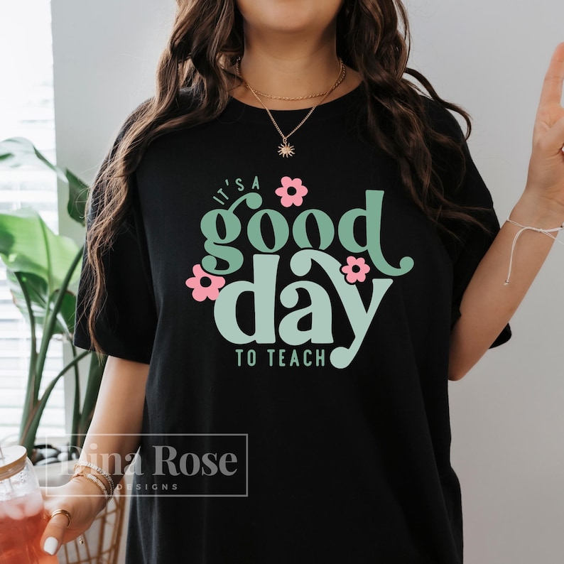 May include: Black t-shirt with a green and white graphic that says "It's a good day to teach" with pink flowers around the text.