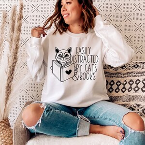 K&ouml;nnte beinhalten: Wei&szlig;er Pullover mit einer schwarz-wei&szlig;en Illustration einer Katze, die ein Buch liest. Der Text auf dem Pullover lautet "Easily Distracted by Cats & Books".