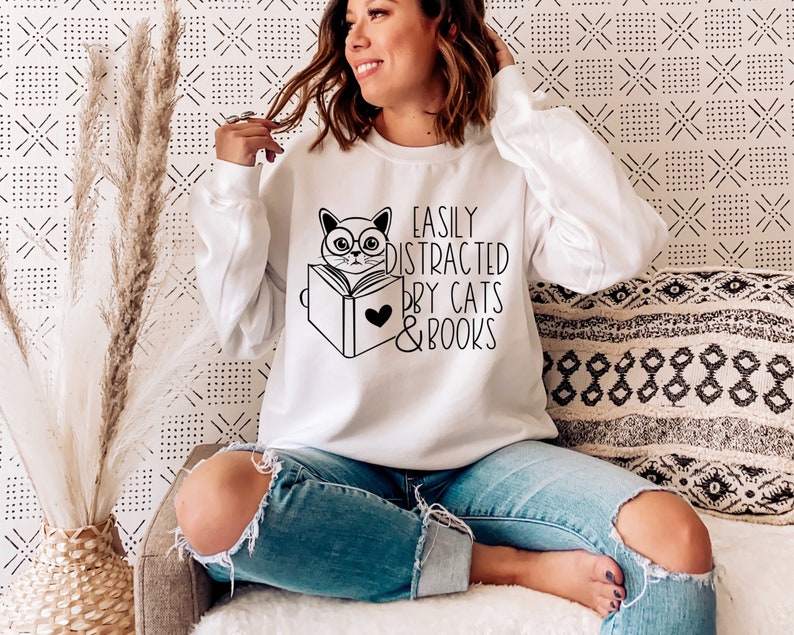 K&ouml;nnte beinhalten: Wei&szlig;er Pullover mit einer schwarz-wei&szlig;en Illustration einer Katze, die ein Buch liest. Der Text auf dem Pullover lautet "Easily Distracted by Cats & Books".