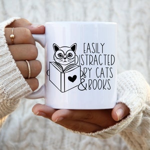 K&ouml;nnte beinhalten: Eine wei&szlig;e Keramiktasse mit einer Schwarzwei&szlig;illustration einer Katze mit Brille, die ein Buch liest. Der Text auf der Tasse lautet "Easily Distracted by Cats & Books".