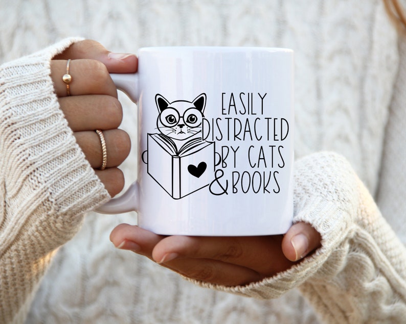 K&ouml;nnte beinhalten: Eine wei&szlig;e Keramiktasse mit einer Schwarzwei&szlig;illustration einer Katze mit Brille, die ein Buch liest. Der Text auf der Tasse lautet "Easily Distracted by Cats & Books".