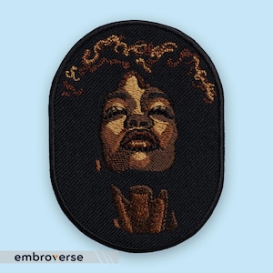 Peut inclure: Écusson brodé de forme ovale représentant un portrait détaillé d'une personne à la peau foncée et aux cheveux bruns. Le visage est rendu dans des tons bruns et beiges, avec les cheveux dans un brun plus clair. Le patch a une bordure noire et le mot "embroverse" est en bas.