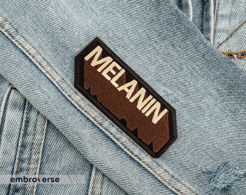 Peut inclure: Un &eacute;cusson brod&eacute; marron et noir avec le mot "MELANIN" en lettres blanches sur une veste en jean.