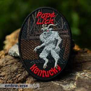 May include: Embroidered patch with a white, furry creature with red eyes and horns. The creature is standing in a dark forest with the text "Pope Lick Kentucky" in red and black.