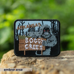 May include: Embroidered patch featuring a gray Bigfoot holding a sign that reads "Boggy Creek" with "Arkansas" above. The patch has a black border and a light blue background with trees. The Bigfoot has red eyes.