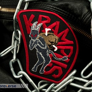 Puede incluir: Un parche bordado negro y rojo con un demonio negro que lleva un saco marrón. El texto "KRAMPUS" está escrito en rojo alrededor del demonio.