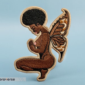 Puede incluir: Parche bordado de una mujer de piel morena con afro, vistiendo un vestido marrón y alas de mariposa. La mujer está arrodillada con las manos juntas frente a ella.