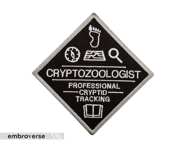 May include: Black and white embroidered patch in a diamond shape. The patch reads "Cryptozoologist" and "Professional Cryptid Tracking". It features a compass, footprint, and magnifying glass graphic.