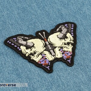 以下が含まれることがあります： 頭蓋骨のデザインが施された蝶の黒い刺繍パッチ。蝶は紫と白のアクセントが付いています。