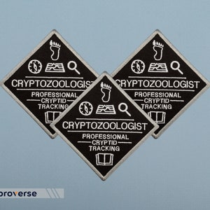 May include: Three black diamond-shaped embroidered patches with white text that reads "Cryptozoologist - Professional Cryptid Tracking". Each patch features a compass, a footprint, and a magnifying glass.