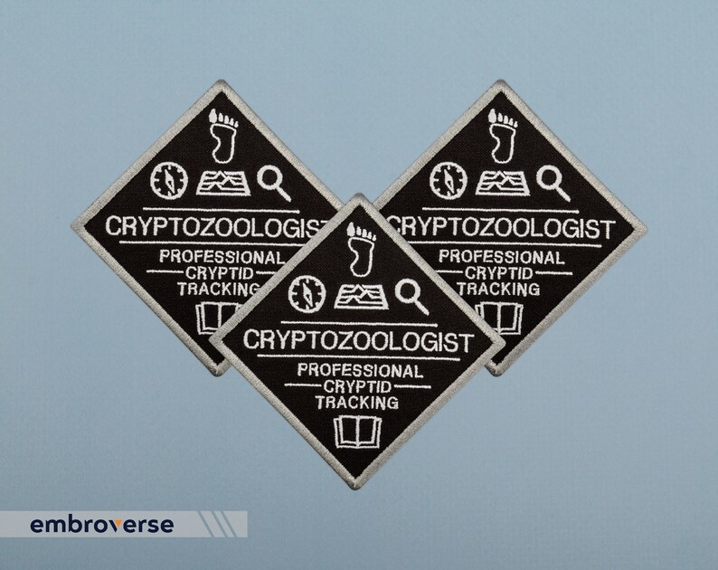 May include: Three black diamond-shaped embroidered patches with white text that reads "Cryptozoologist - Professional Cryptid Tracking". Each patch features a compass, a footprint, and a magnifying glass.