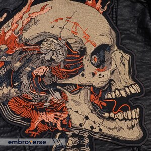 May include: Embroidered patch featuring a detailed skull design with exposed internal mechanics and fiery orange accents. The skull is beige with black and orange details, set against a black background. The text "embroverse" is visible.