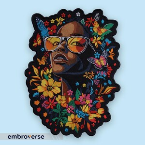 Puede incluir: Parche bordado que representa a una mujer con gafas de sol, rodeada de flores y mariposas coloridas. El parche tiene un borde negro y la palabra "embroverse" está impresa en la parte inferior.