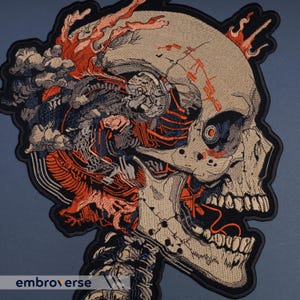 May include: Embroidered patch featuring a detailed, stylized skull with exposed internal components. The design incorporates shades of beige, black, orange, and gray, with a dark border. The text "embroverse" is visible at the bottom.