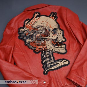 May include: A red leather jacket with a large embroidered skull patch on the back. The patch features a detailed design with beige, black, and orange accents. The jacket has a classic collar and a button closure. The text "embroverse" is visible at the bottom.