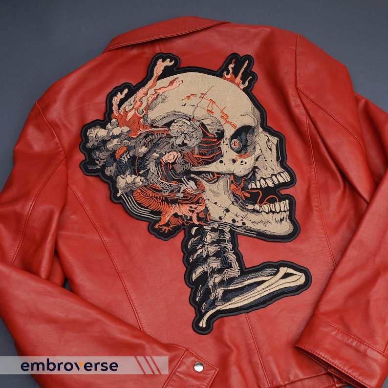 May include: A red leather jacket with a large embroidered skull patch on the back. The patch features a detailed design with beige, black, and orange accents. The jacket has a classic collar and a button closure. The text "embroverse" is visible at the bottom.