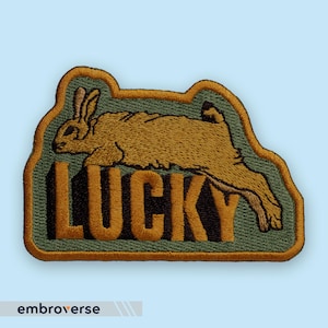 Peut inclure: Écusson brodé avec un lapin courant marron et beige, et le mot "LUCKY" en orange foncé. L'écusson a un fond vert et une bordure dorée. Le nom de la marque "embroverse" est en bas.