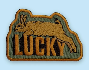 May include: Embroidered patch featuring a running rabbit in shades of brown and tan, with the word "LUCKY" in bold, dark orange letters. The patch has a green background and a gold border. The brand name "embroverse" is at the bottom.