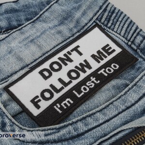 May include: A close-up of a pair of blue denim jeans with a black and white patch that reads "Don't Follow Me I'm Lost Too".