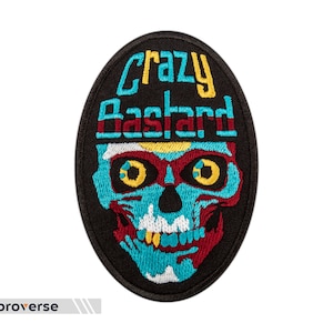 Puede incluir: Un parche bordado en blanco y negro con un diseño de calavera y huesos cruzados. La calavera es azul y roja con ojos amarillos. El texto "Crazy Bastard" está escrito en amarillo y azul sobre la calavera.