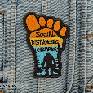 Puede incluir: Parche bordado con forma de huella de Bigfoot, con el texto "Social Distancing Champion". El parche presenta colores naranja, amarillo, azul y negro, y una silueta de Bigfoot.