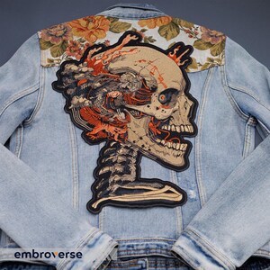 May include: Light blue denim jacket with a large skull patch on the back. The patch features a detailed illustration of a skull with orange and brown accents. The jacket's upper back has a floral pattern.