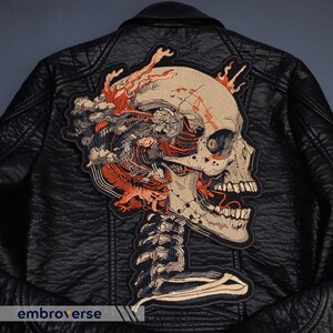 May include: Black leather jacket with a large embroidered patch featuring a detailed skull design. The patch incorporates shades of beige, orange, and blue, with intricate anatomical details and a skeletal structure. The jacket has a classic biker style.