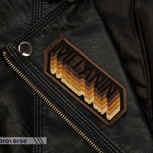 Peut inclure: Une veste en cuir marron avec un patch brod&eacute; marron et beige qui dit "MELANIN" dans une police r&eacute;tro.