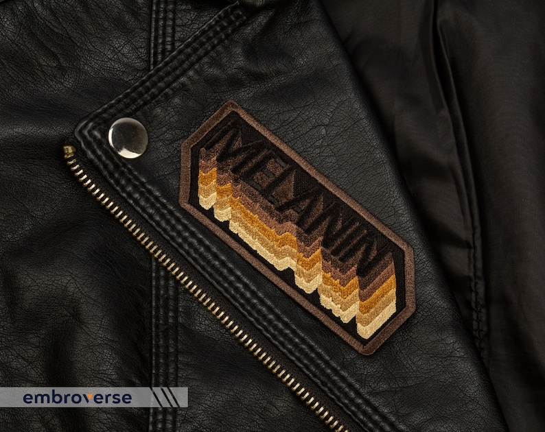 Peut inclure: Une veste en cuir marron avec un patch brod&eacute; marron et beige qui dit "MELANIN" dans une police r&eacute;tro.