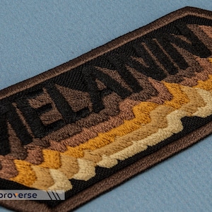 Peut inclure: Patch brod&eacute; avec le mot "MELANIN" en fil noir sur un fond brun. Le fond pr&eacute;sente un design en couches de fil brun, beige et or.