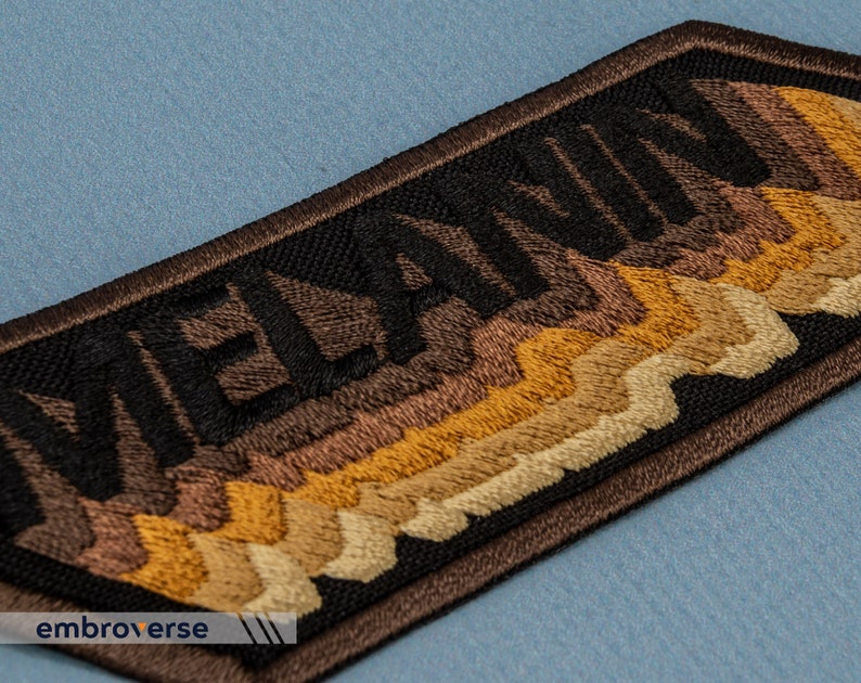 Peut inclure: Patch brod&eacute; avec le mot "MELANIN" en fil noir sur un fond brun. Le fond pr&eacute;sente un design en couches de fil brun, beige et or.