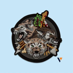 May include: A circular embroidered patch featuring three snarling wolves with green eyes. The design includes a central wolf head flanked by two others, surrounded by crossed arrows, a bone, a stone-headed axe, and a small green leafy branch.