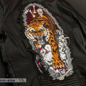 May include: A black leather jacket featuring an embroidered patch of a tiger with a skull and bones design. The tiger is orange and black with white stripes. The patch is surrounded by a barbed wire design.