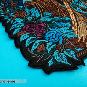 Puede incluir: Un parche bordado negro con un dise&ntilde;o detallado de un arreglo floral con flores azules, moradas, marrones y verdes. El parche tiene un borde festoneado.