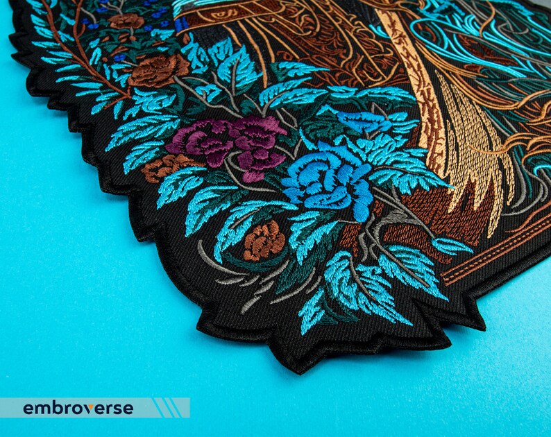 Puede incluir: Un parche bordado negro con un dise&ntilde;o detallado de un arreglo floral con flores azules, moradas, marrones y verdes. El parche tiene un borde festoneado.