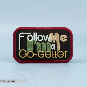 Pode incluir: Um remendo bordado preto com costuras vermelhas ao redor das bordas. O remendo diz "Follow Me I'm a Go-Getter" em fio branco, verde e amarelo.