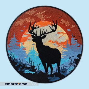 Op de afbeelding: Gestikte patch met een silhouet van een hert dat in een bos staat met een zonsondergang op de achtergrond. Het hert is zwart, het bos is blauw en groen, en de zonsondergang is oranje en rood.