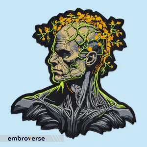 Könnte beinhalten: Ein schwarzer und grüner gestickter Patch, der einen menschlichen Kopf mit einem schädelartigen Gesicht und einer Krone aus gelben Blättern zeigt. Der Kopf ist von grünen Ranken umgeben und das Gesicht hat eine grüne und gelbe Skelettstruktur.