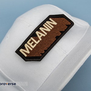 Peut inclure: Casquette de baseball blanche avec un patch brod&eacute; marron et noir qui dit "MELANIN" en lettres blanches.