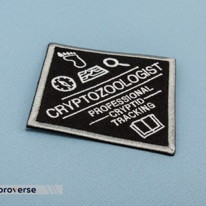 May include: Black and white embroidered patch with the text "Cryptozoologist Professional Cryptid Tracking" and various symbols including a compass, footprint, magnifying glass, and an open book.